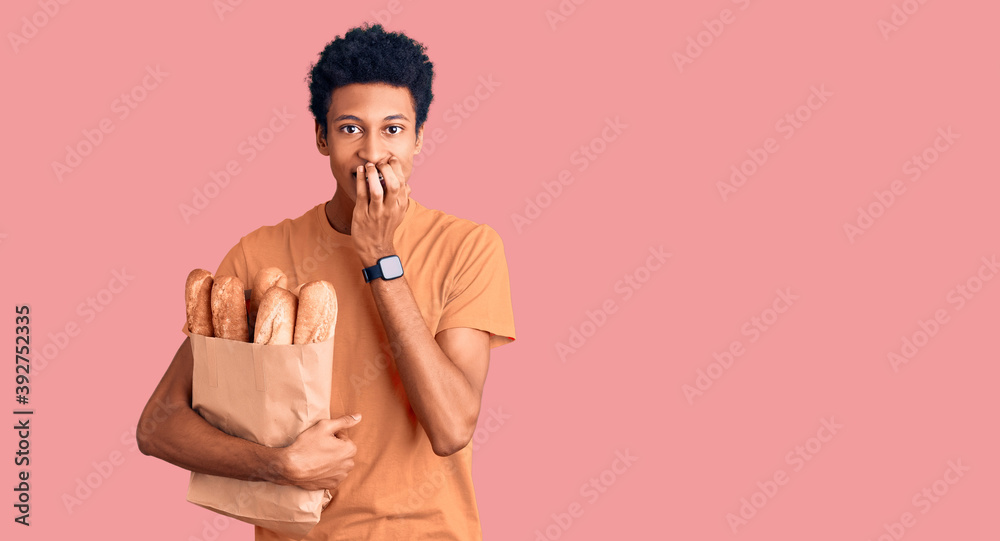 Fototapeta premium Young african american man holding paper bag with bread laughing and embarrassed giggle covering mouth with hands, gossip and scandal concept