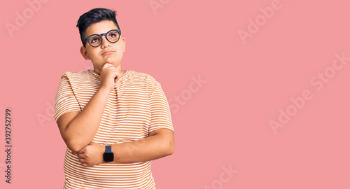 Little boy kid wearing casual clothes and glasses with hand on chin thinking about question, pensive expression. smiling with thoughtful face. doubt concept.