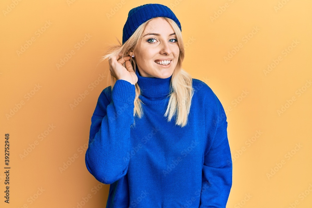 Young caucasian woman wearing wool winter sweater and cap smiling with hand over ear listening and hearing to rumor or gossip. deafness concept. Young caucasian woman wearing wool winter sweater and cap smiling with hand over ear listening and hearing to rumor or gossip. deafness concept.