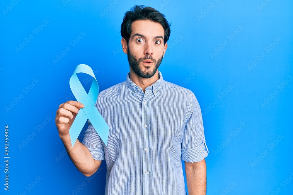 Young hispanic man holding blue ribbon scared and amazed with open mouth for surprise, disbelief face Young hispanic man holding blue ribbon scared and amazed with open mouth for surprise, disbelief face