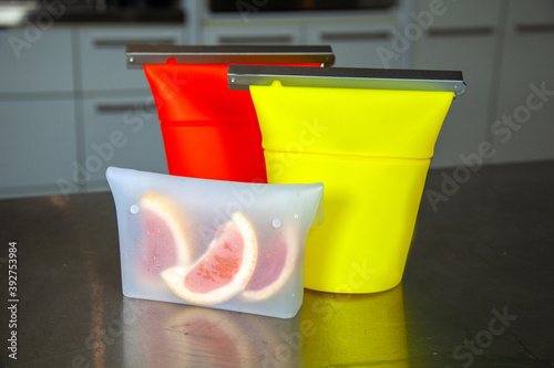 Three zero-waste food-grade silicone food bags to hold snacks or dried goods to replace plastic zip-lock style bags. A red and yellow bag stand beside a clear snack bag with grapefruit. photo