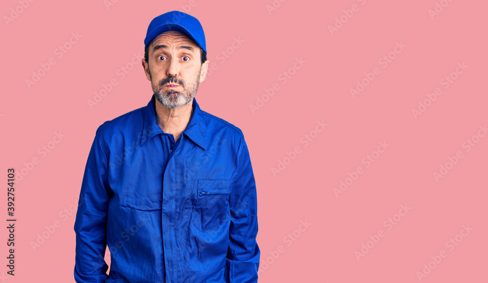 Middle age handsome man wearing mechanic uniform puffing cheeks with funny face. mouth inflated with air, crazy expression. Middle age handsome man wearing mechanic uniform puffing cheeks with funny face. mouth inflated with air, crazy expression.