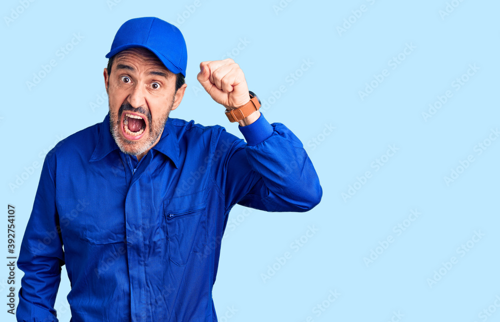 Middle age handsome man wearing mechanic uniform angry and mad raising fist frustrated and furious while shouting with anger. rage and aggressive concept.