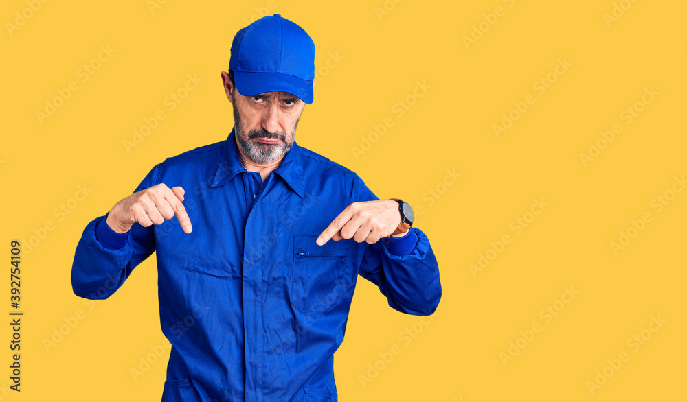 Middle age handsome man wearing mechanic uniform pointing down looking sad and upset, indicating direction with fingers, unhappy and depressed.
