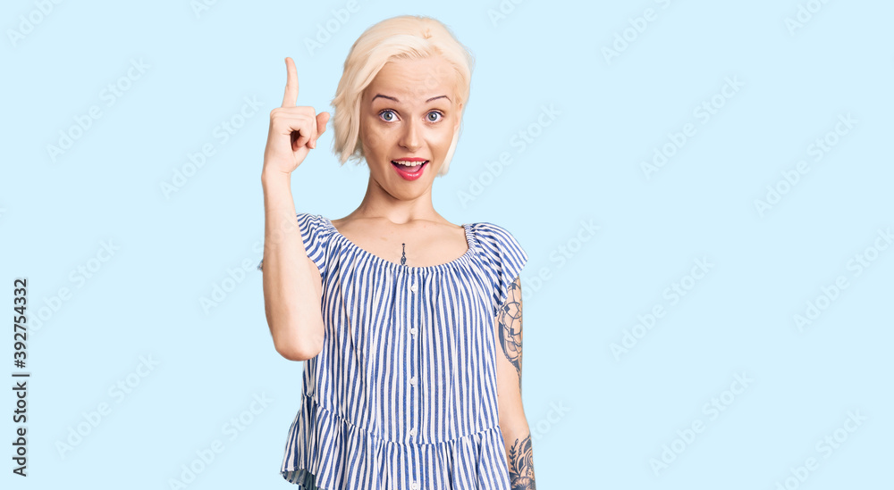 Young blonde woman with tattoo wearing casual clothes pointing finger up with successful idea. exited and happy. number one.