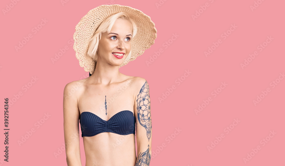 Young blonde woman with tattoo wearing bikini and summer hat looking away to side with smile on face, natural expression. laughing confident. Young blonde woman with tattoo wearing bikini and summer hat looking away to side with smile on face, natural expression. laughing confident.