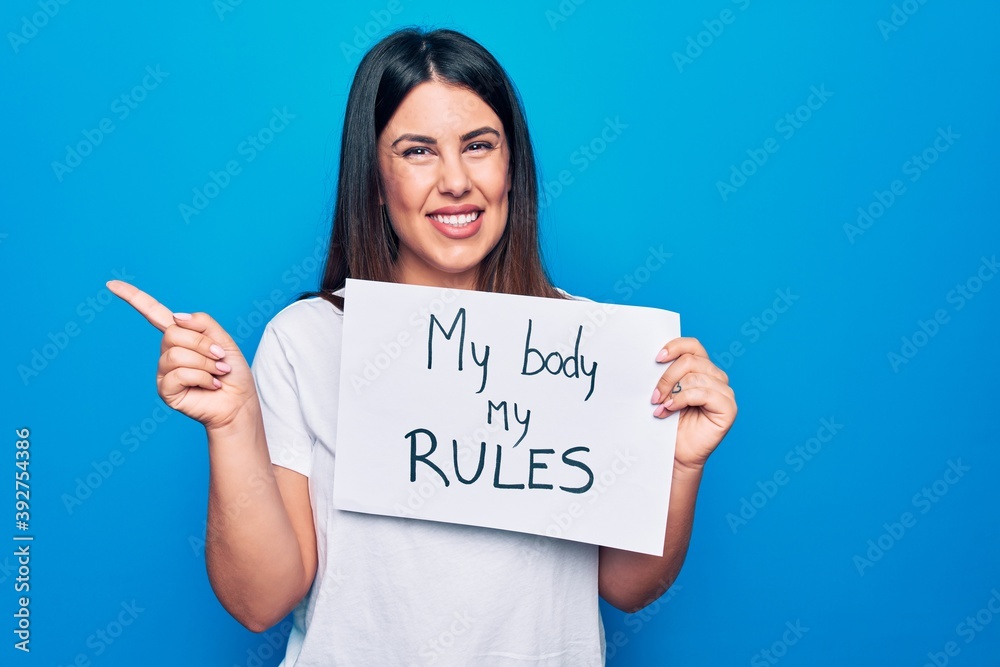 Young beautiful woman asking for women rights holding paper with my body my rules message smiling happy pointing with hand and finger to the side Young beautiful woman asking for women rights holding paper with my body my rules message smiling happy pointing with hand and finger to the side