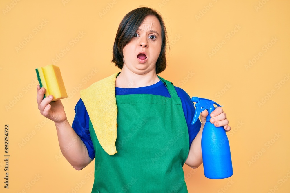Brunette woman with down syndrome wearing apron holding scourer in shock face, looking skeptical and sarcastic, surprised with open mouth Brunette woman with down syndrome wearing apron holding scourer in shock face, looking skeptical and sarcastic, surprised with open mouth