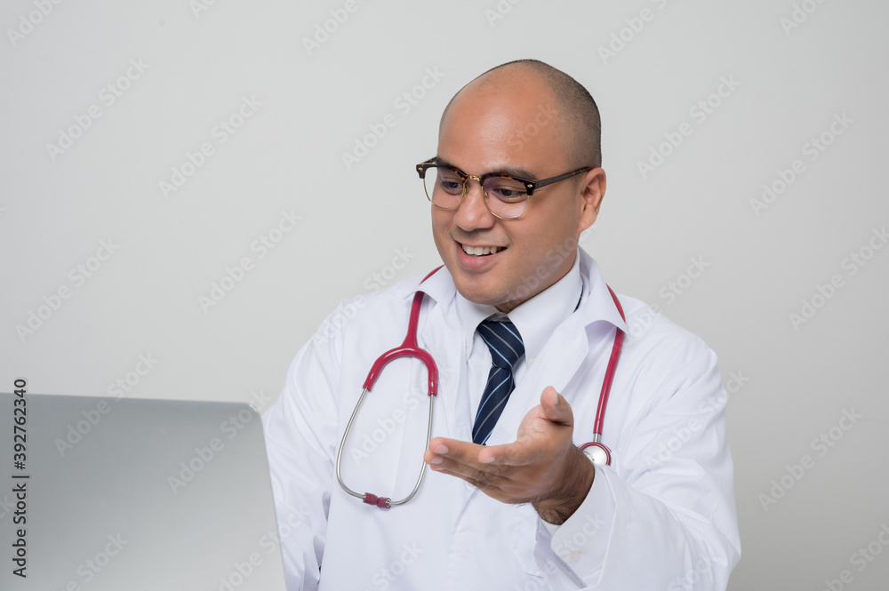© Chanakon - A portrait of Asian doctor put on stethoscope look into laptop and talk sitting at the desk isolated on white background.