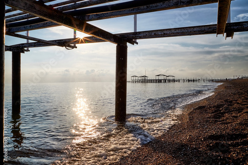 View from sand beach to water of sea, waves and pier in a nice day or evening with blues sky, bright sun and white clouds. The concept of a holiday on the sea or ocean in the South. © keleny