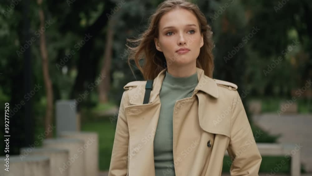 Attractive student girl dressed in trench coat going home after college through cozy city park Attractive student girl dressed in trench coat going home after college through cozy city park