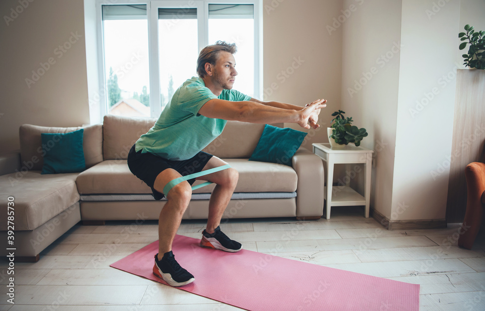 Caucasian man is doing fitness at home using special bands on the floor while squatting