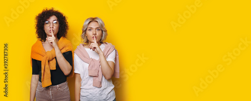 Two caucasian women are gesturing the silence sign on a yellow wall with free space looking seriously at camera photo