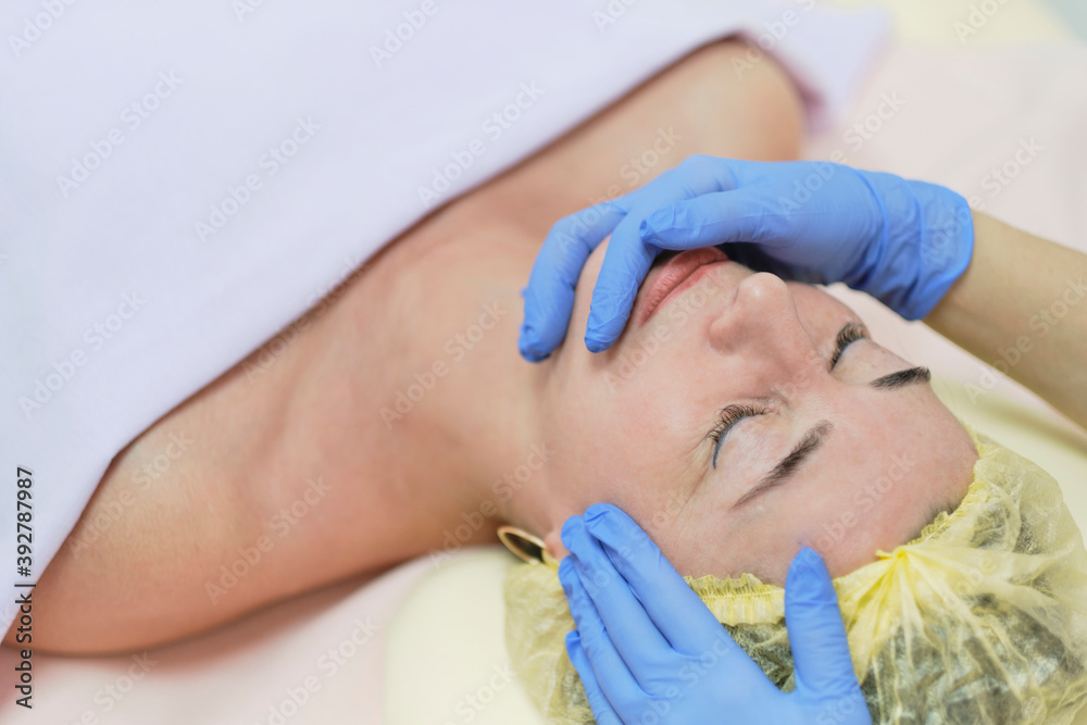 Close up portrait of charming woman with closed eyes enjoying skincare procedure. Masseuse arms touching client chin and nose. Anti-aging effect