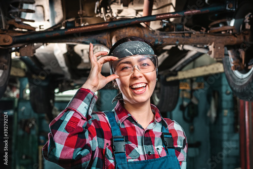 Portrait of young happy repairwoman, in a uniform and glasses poses standing under a car on a lift. Indoors garage