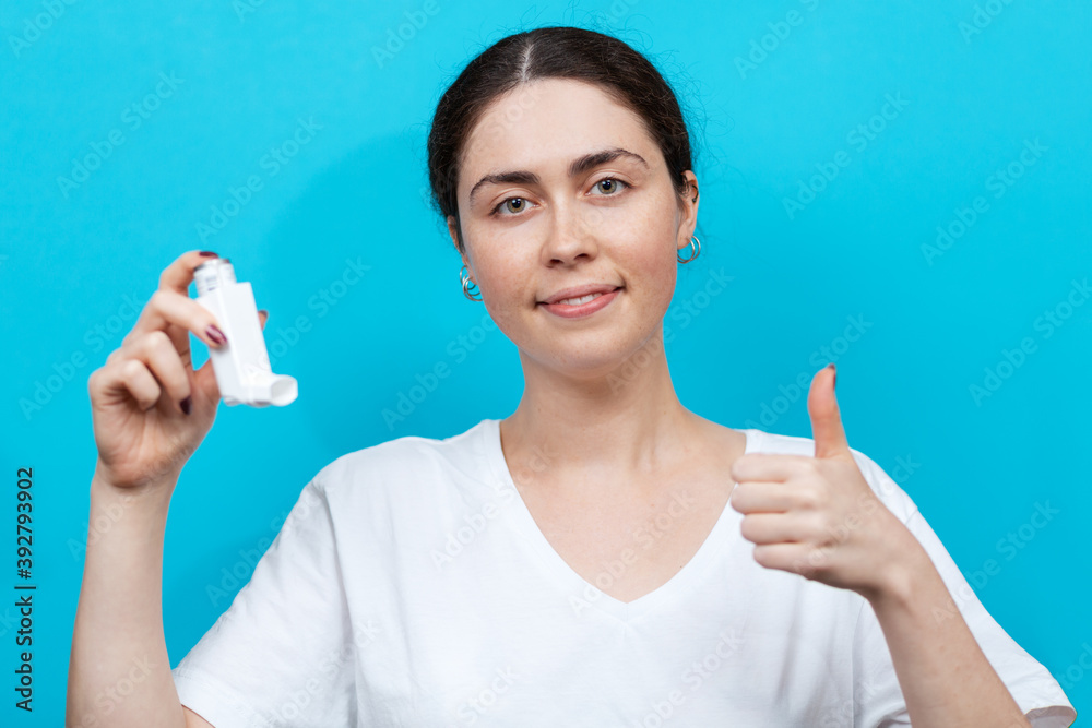 Obraz premium Portrait of a female smiling doctor holding an inhaler in her hand and giving a thumbs up. Blue background