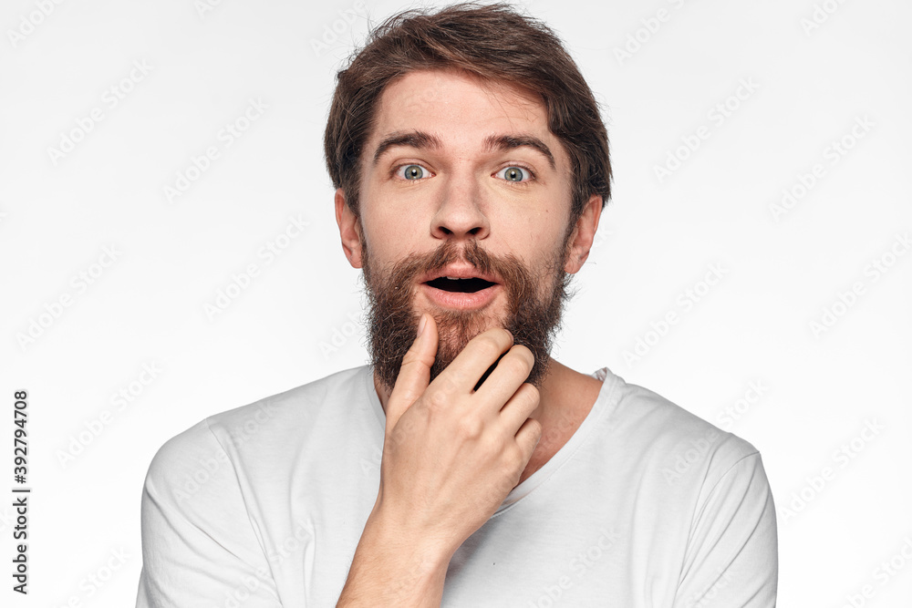 cheerful bearded man in a white t-shirt emotions gestures with his hands light background studio