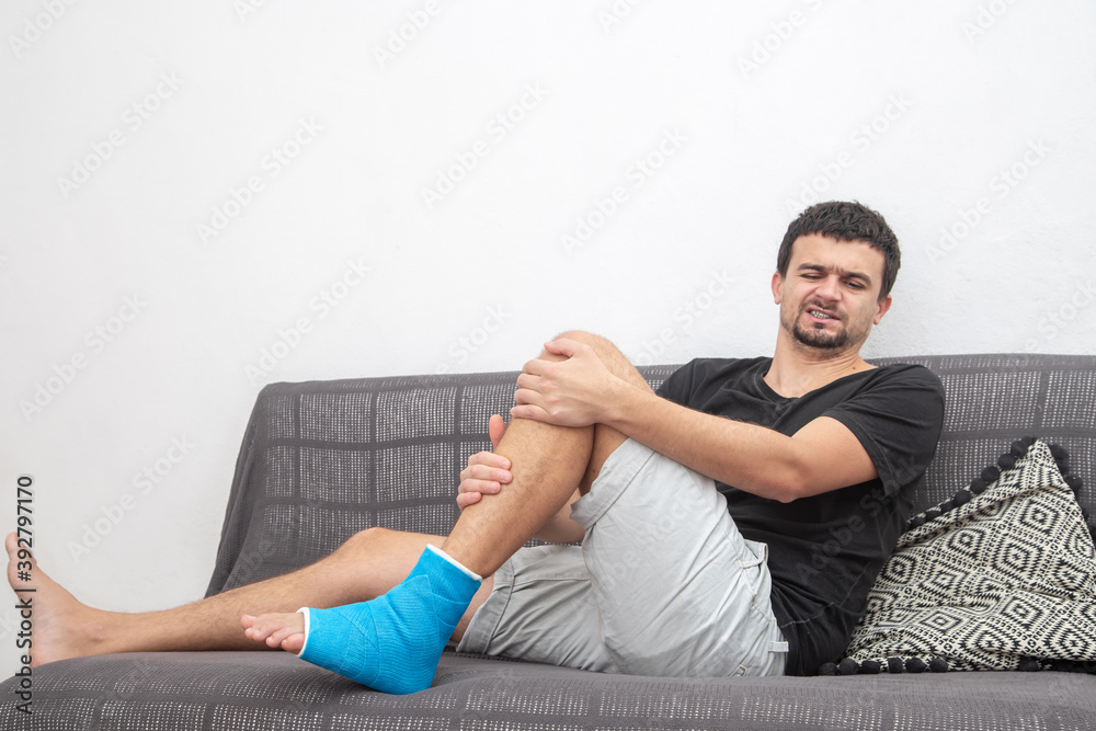 A young man with a broken leg in a cast lies on a sofa and writhes in pain