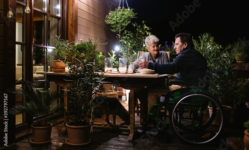 Senior couple in wheelchair having dinner in the evening on terrace, drinking wine.