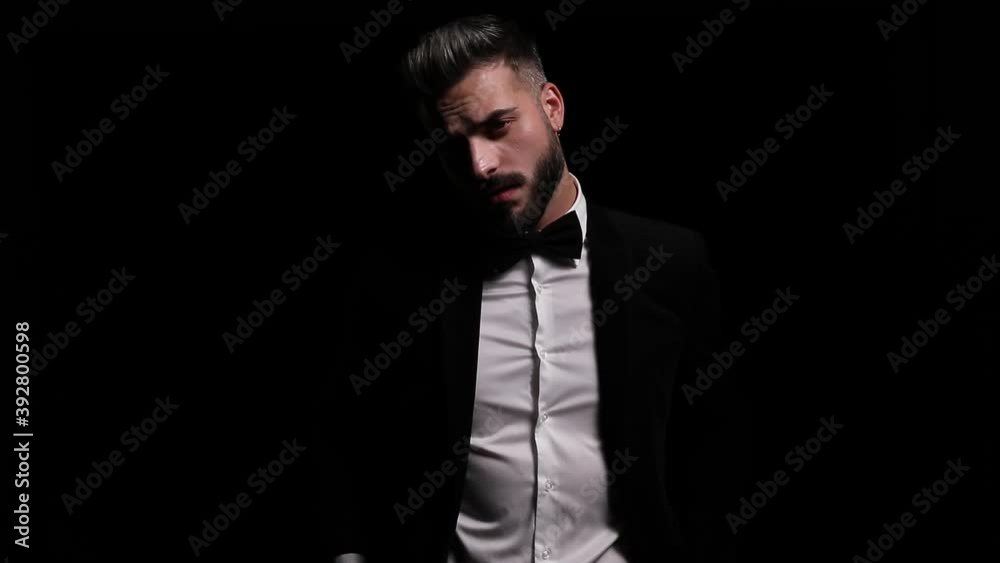 mysterious young man arranging bowtie and black tuxedo, moving in a side view position and posing, smiling, snapping fingers and leaving on black background in studio
