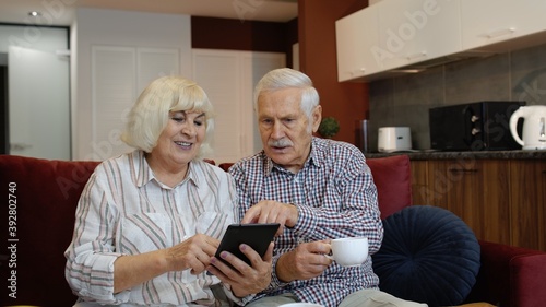 Happy smiling senior couple with tablet pc computer at home. Cheerful retired husband and wife hugging and laughing using digital tablet resting on sofa in cozy living room