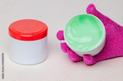 Closeup of a closed and an open container with cooling gel (horse balm) for sports injuries on a light background. The open container is held by a pink massage glove. Copy space. Macro. photo