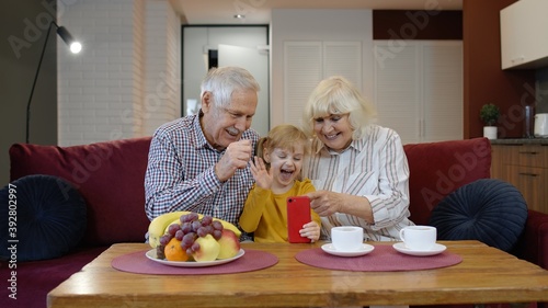 Caucasian grandfather, grandmother and small granddaughter making video call online on mobile phone at home. Little child kid girl with grandparents talking by internet webcam chat app on smartphone