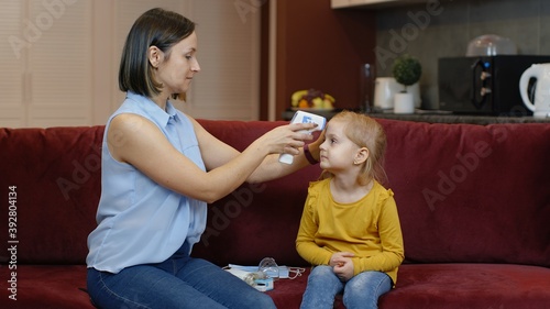 Mother measures temperature with contactless digital thermometer of her little sick daughter child girl at home. Woman with non-contact infrared thermometer. Coronavirus  Covid-19 quarantine lockdown