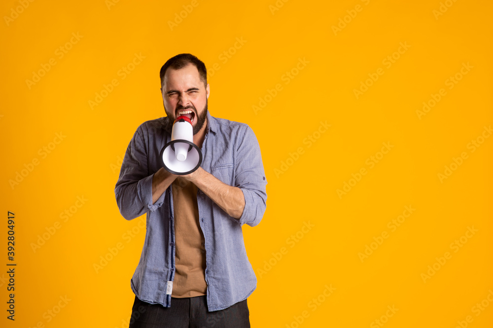 a bearded man in a casual shirt with a loudspeaker in his hands shouts into a megaphone on a yellow background. a bearded man in a casual shirt with a loudspeaker in his hands shouts into a megaphone on a yellow background.