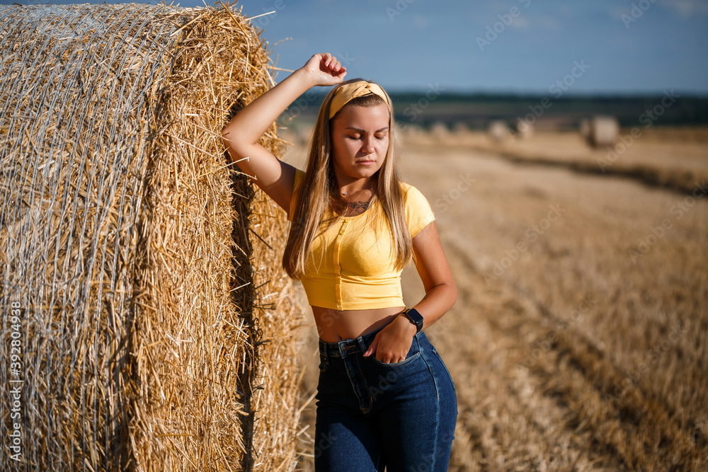 A young beautiful girl in a field stands near a sheaf. Vacation in the village. Young woman in yellow top and jeans A young beautiful girl in a field stands near a sheaf. Vacation in the village. Young woman in yellow top and jeans
