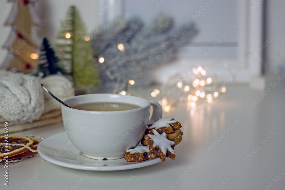 Homemade christmas tea party background. White mug of tea with asterisk cookies, dried oranges, cinnamon sticks on a white table, surrounded by new decor and garlands. Cozy winter, hygge atmosphere Homemade christmas tea party background. White mug of tea with asterisk cookies, dried oranges, cinnamon sticks on a white table, surrounded by new decor and garlands. Cozy winter, hygge atmosphere