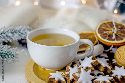 Homemade christmas tea party background. White mug of tea with asterisk cookies, dried oranges, cinnamon sticks on a white table, surrounded by new decor and garlands. Cozy winter, hygge atmosphere