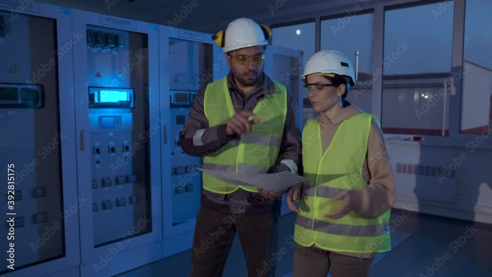 Professional electric power station engineers in reflective vests walk past lit blue special sensor control cabinets in room closeup. Night shift of the station staff. Professional electric power station engineers in reflective vests walk past lit blue special sensor control cabinets in room closeup. Night shift of the station staff.