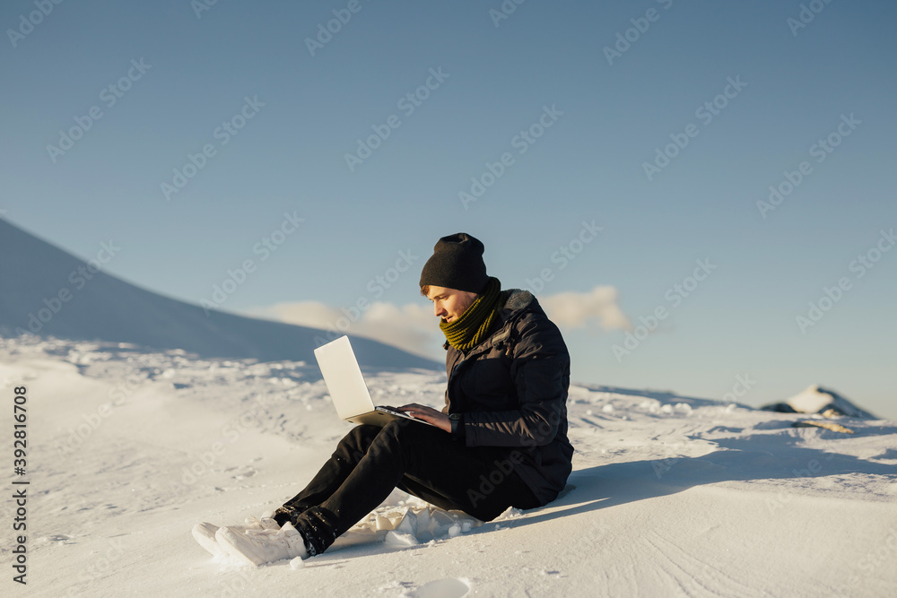 Tourist man on the snowy mountain using laptop on mountain peak with blue sky on the background. Copy space.