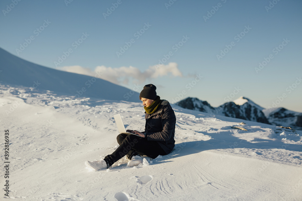 Man sitting on the snowy mountain and using laptop on mountain peak with blue sky on the background. Copy space.