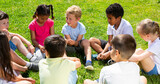 Group of happy kids resting on grass together and chatting in park at summer day