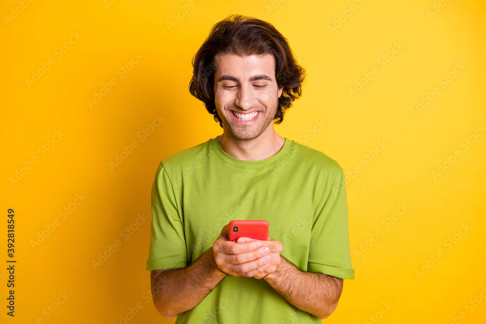 Photo of cheerful young guy hold telephone look screen wear green t-shirt isolated yellow color background Photo of cheerful young guy hold telephone look screen wear green t-shirt isolated yellow color background