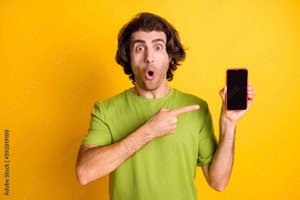 Photo of guy hold phone indicate forefinger screen empty space open mouth wear green t-shirt isolated yellow color background Photo of guy hold phone indicate forefinger screen empty space open mouth wear green t-shirt isolated yellow color background