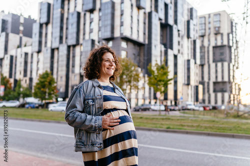 Active mom. Portrait of happy young business pregnant woman looking at camera while standing on the street with document folder. Smiling future mom working on her late pregnancy period