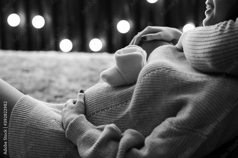 Black and white cropped image of beautiful happy pregnant woman and her husband holding baby shoes and hugging the tummy.