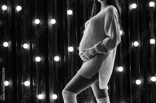 Black and white cropped image of pregnant woman in white sweater touching her belly at  black background with lights. Future mother is waiting her baby.