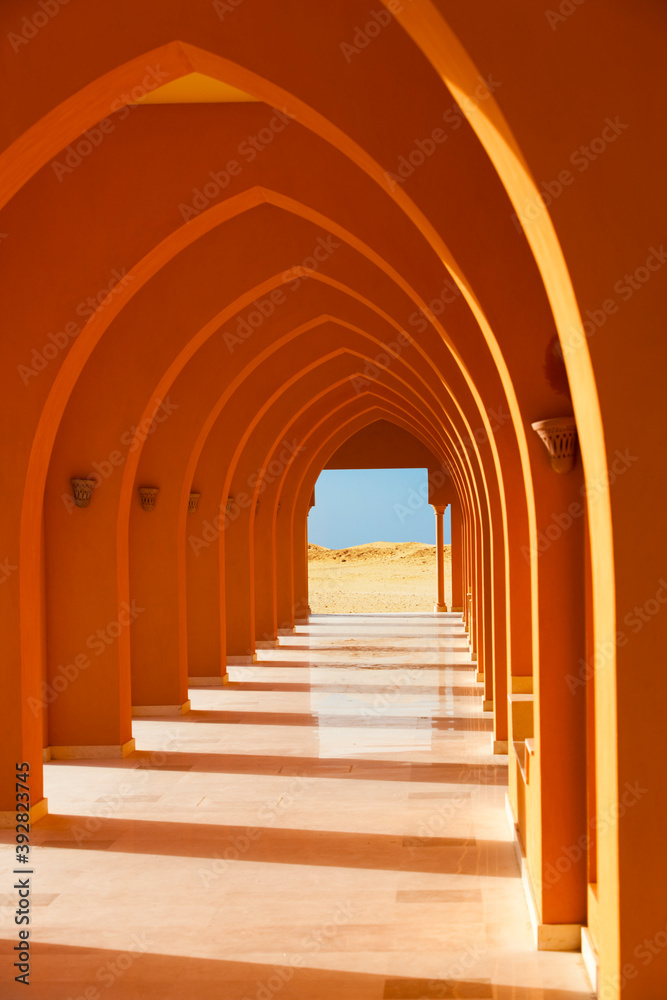 Colorful orange arched hallway passage with columns leading to a desert on a sunny day.