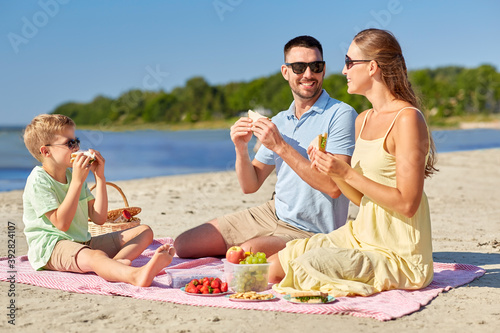 family, leisure and people concept - happy mother, father and little son having picnic on summer beach and eating sandwiches