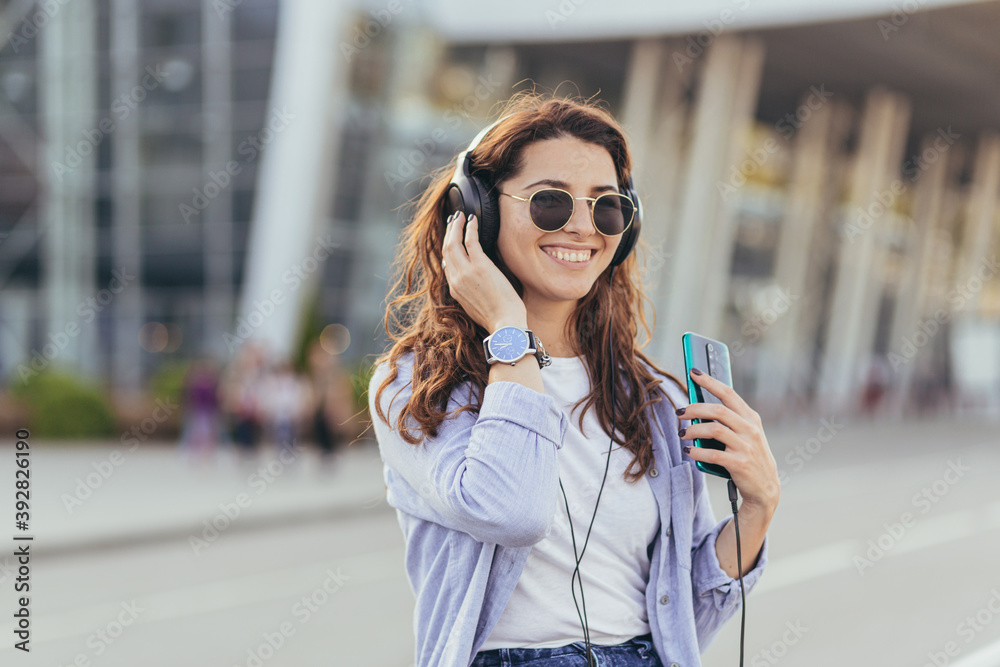 Young pretty student girl, waiting for a taxi car, and listening to music from the phone on big headphones