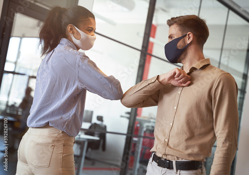 Two young diverse business colleagues wearing face protective masks bumping elbows, greeting each other while working during covid 19 quarantine photo
