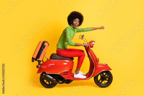 Photo portrait of excited brunette african american woman on motorcycle with orange suitcase arm fist forward isolated on bright yellow colored background © deagreez
