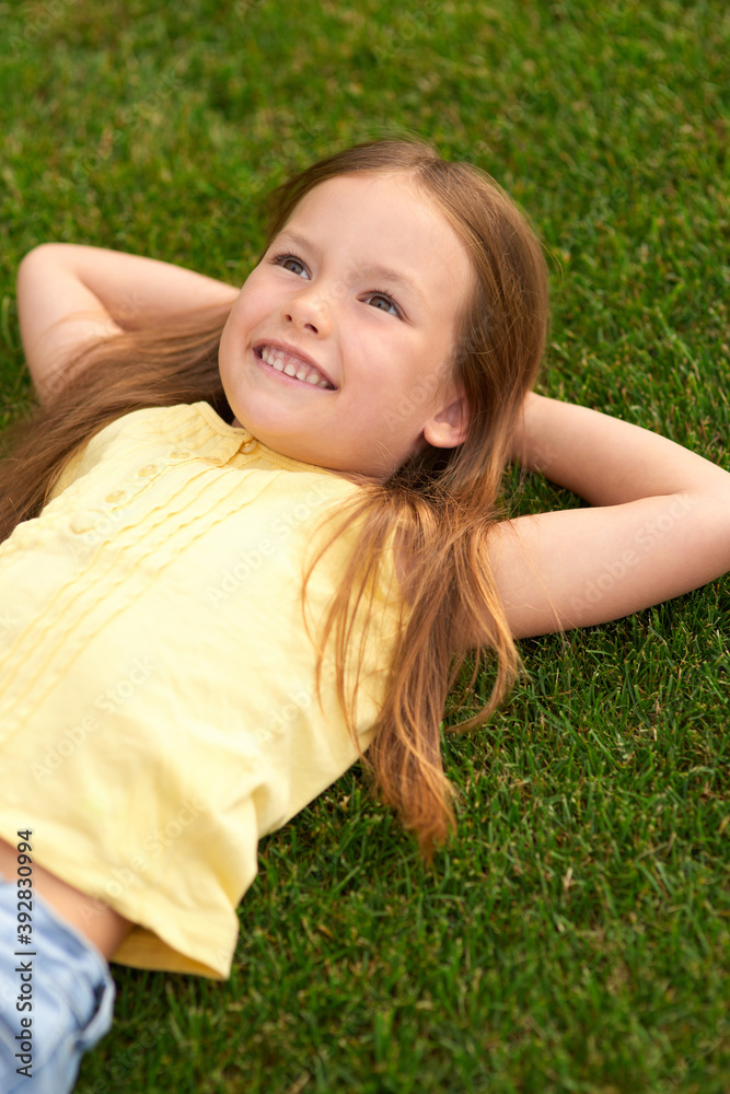 Dreaming... Portrait of joyful little girl in casual clothes lying on the green lawn, looking up and smiling while visiting park on a sunny summer day Dreaming... Portrait of joyful little girl in casual clothes lying on the green lawn, looking up and smiling while visiting park on a sunny summer day