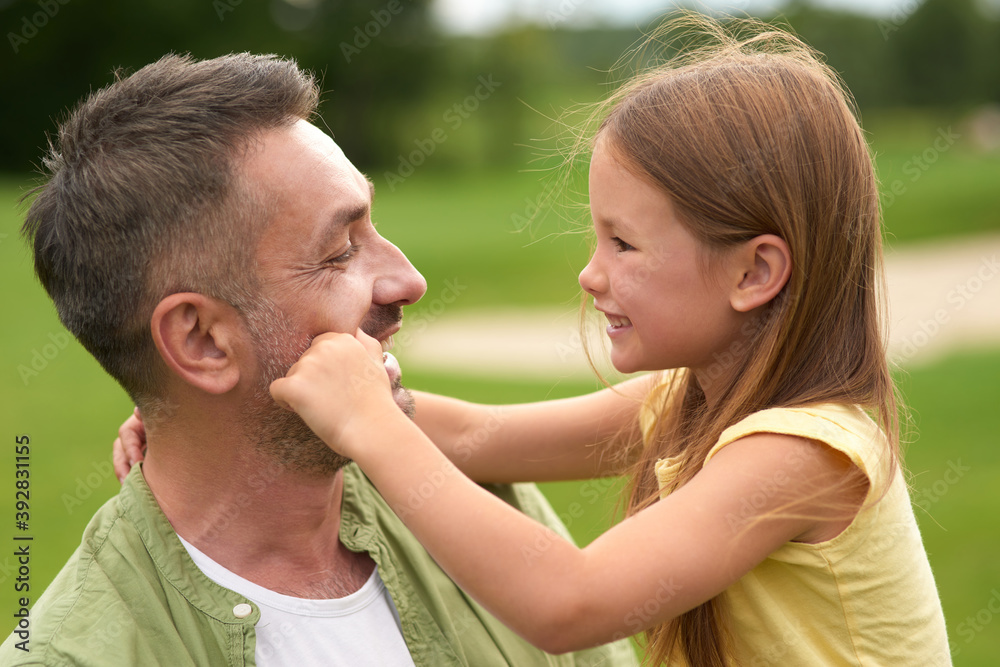 Close up of adorable little girl having fun with her loving daddy, touching his face while he is holding her and smiling, standing in the green park Close up of adorable little girl having fun with her loving daddy, touching his face while he is holding her and smiling, standing in the green park