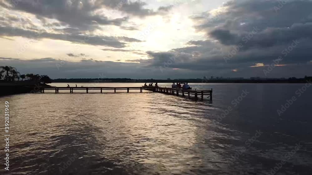 Zoom out mostrando o céu parcialmente nublado e um pier com pessoas sentadas Zoom out mostrando o céu parcialmente nublado e um pier com pessoas sentadas