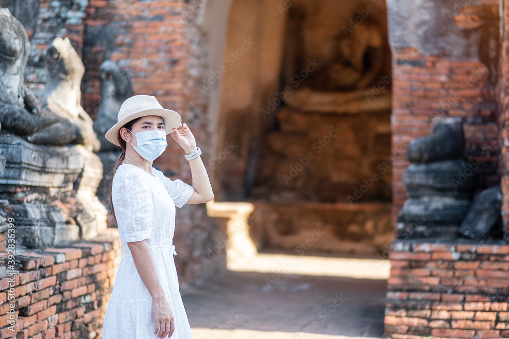 Tourist Woman in white dress wearing surgical face mask, protection COVID-19 pandemic during visiting in Wat Chaiwatthanaram temple in Ayutthaya. new normal, safety travel and Thailand travel concept